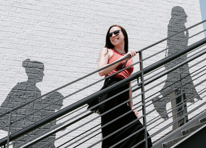 brunette girl with sunglasses on stairs smiling over shoulder, peoples shadows in front of and behind her on white brick wall
